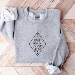 Puede incluir: Una sudadera gris con un gr&aacute;fico negro del s&iacute;mbolo del zod&iacute;aco Piscis, dos peces nadando dentro de un diamante con estrellas y el s&iacute;mbolo de Piscis.