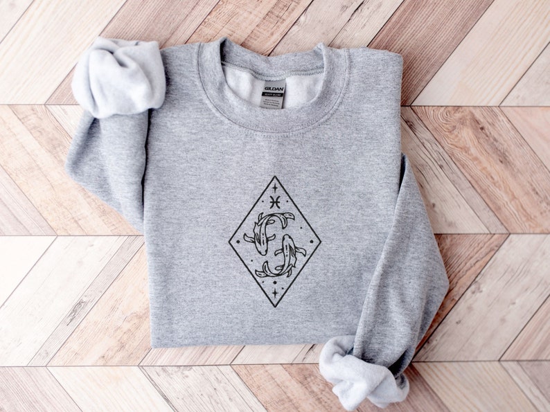 Puede incluir: Una sudadera gris con un gr&aacute;fico negro del s&iacute;mbolo del zod&iacute;aco Piscis, dos peces nadando dentro de un diamante con estrellas y el s&iacute;mbolo de Piscis.