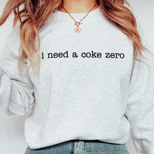 以下が含まれることがあります： 前面に「i need a coke zero」という文字がプリントされたグレーのスウェットシャツです。