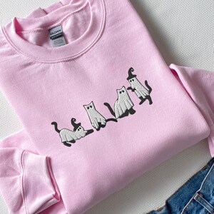 May include: A pink sweatshirt with an embroidered design of four white cats dressed as ghosts wearing witch hats. The cats are in a line, facing the right side of the image.