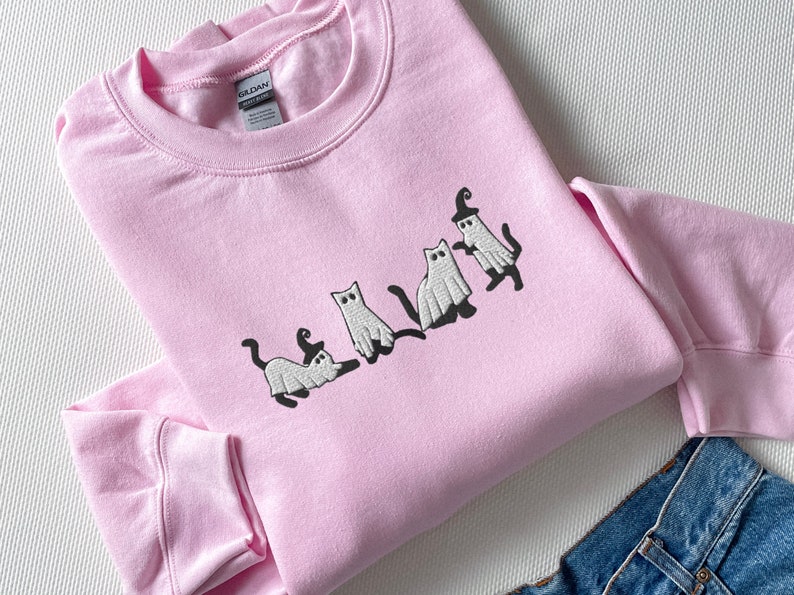May include: A pink sweatshirt with an embroidered design of four white cats dressed as ghosts wearing witch hats. The cats are in a line, facing the right side of the image.