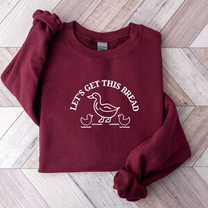 May include: A maroon sweatshirt with a white graphic of a duck and ducklings with the text "Let's get this bread".