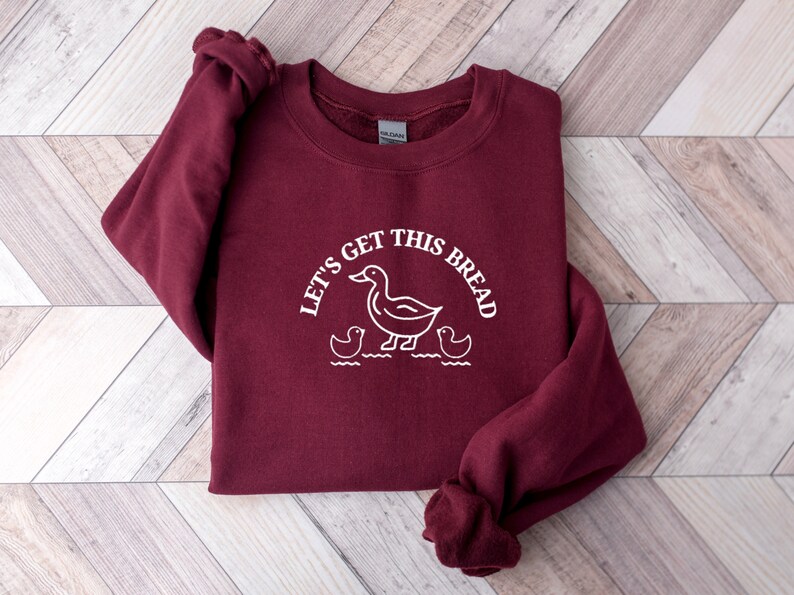 May include: A maroon sweatshirt with a white graphic of a duck and ducklings with the text "Let's get this bread".