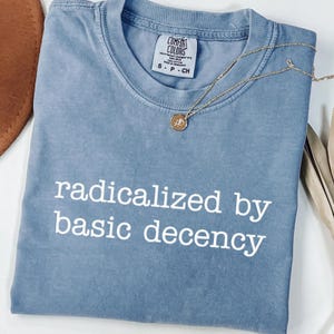 Könnte beinhalten: Ein blaues T-Shirt mit dem weißen Schriftzug "radicalized by basic decency". Eine goldene Halskette mit einem kleinen Anhänger liegt auf dem gefalteten Shirt. Das Shirt hat ein Comfort Colors-Etikett.