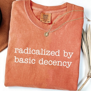 Puede incluir: Una camiseta naranja Comfort Colors con la frase "radicalized by basic decency" en blanco. Un collar dorado con un colgante está drapeado sobre la camiseta. La camiseta está doblada cuidadosamente sobre una superficie blanca.