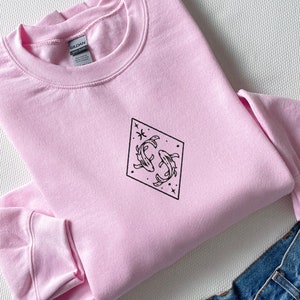 Puede incluir: Una sudadera rosa con un gr&aacute;fico negro de dos peces nadando en un diamante con estrellas.