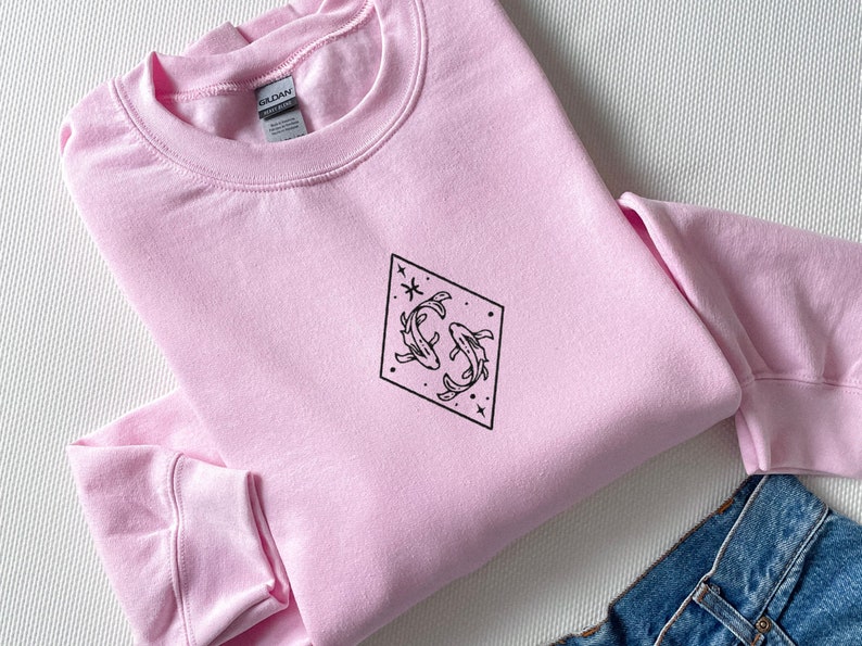 Puede incluir: Una sudadera rosa con un gr&aacute;fico negro de dos peces nadando en un diamante con estrellas.