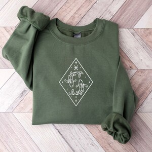Puede incluir: Una sudadera verde con un gr&aacute;fico blanco del s&iacute;mbolo del zod&iacute;aco Piscis, dos peces entrelazados en forma de diamante con estrellas.