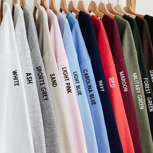 May include: A row of 14 long-sleeved shirts hanging on wooden hangers. The shirts are in various colors, including white, ash, sports grey, sand, light pink, light blue, Carolina blue, navy, red, maroon, military green, forest green, dark chocolate, and black. Each shirt has a color name printed on the sleeve.