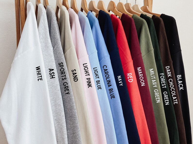 May include: A row of 14 long-sleeved shirts hanging on wooden hangers. The shirts are in various colors, including white, ash, sports grey, sand, light pink, light blue, Carolina blue, navy, red, maroon, military green, forest green, dark chocolate, and black. Each shirt has a color name printed on the sleeve.