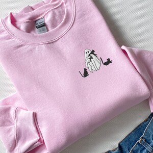 May include: A pink sweatshirt with a white ghost holding a black cat and three black cats sitting below the ghost.