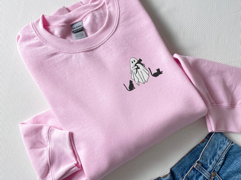 May include: A pink sweatshirt with a white ghost holding a black cat and three black cats sitting below the ghost.