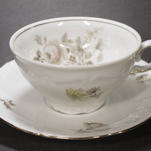 May include: A white teacup and saucer set with a floral pattern in shades of brown and pink. The cup has a gold rim and the saucer has a scalloped edge.