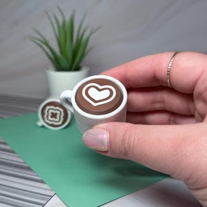 May include: A miniature white ceramic mug with a brown liquid and a white heart design. The mug is being held by a hand.