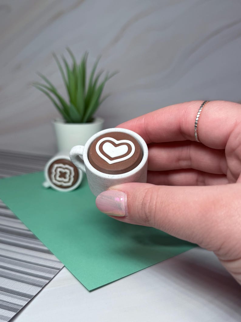 May include: A miniature white ceramic mug with a brown liquid and a white heart design. The mug is being held by a hand.