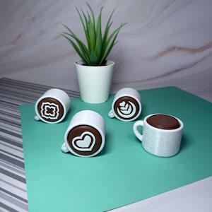 May include: Four white ceramic coffee mugs with brown chocolate-like tops. Each mug has a different white design on the top: a heart, a flower, a swirl, and a geometric pattern. The mugs are on a green surface.
