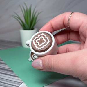 May include: A small white ceramic mug with a brown interior and a white geometric design. The mug is being held in a hand.