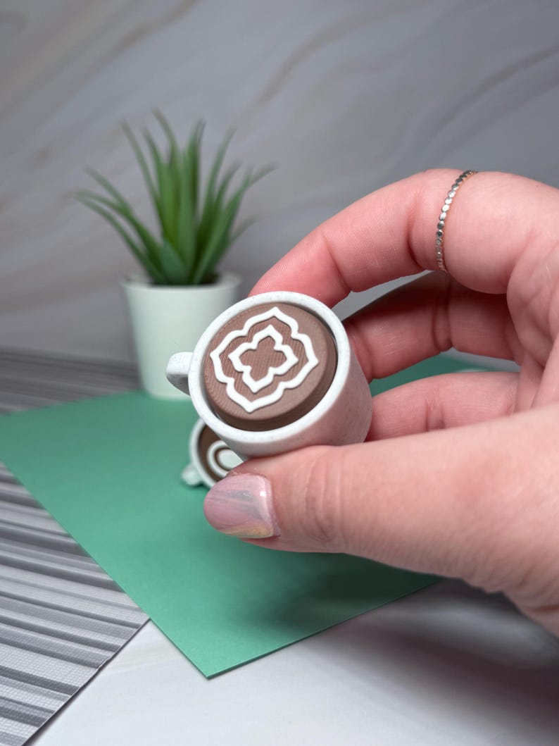 May include: A small white ceramic mug with a brown interior and a white geometric design. The mug is being held in a hand.