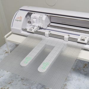 May include: A white electronic cutting machine with a clear, grid-marked cutting mat. The machine has the word "Inspirate" printed on the side. The cutting mat has two cut-out shapes with green accents.