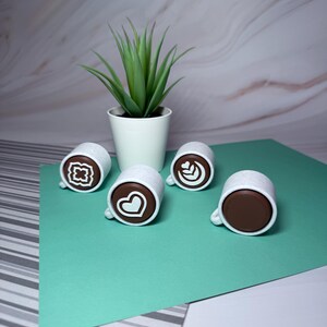 May include: Four white ceramic coffee mugs with brown coffee inside and different latte art designs. The mugs are on a green surface with a white potted succulent plant in the background.
