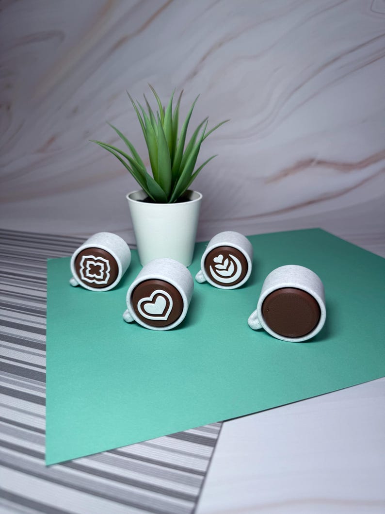 May include: Four white ceramic coffee mugs with brown coffee inside and different latte art designs. The mugs are on a green surface with a white potted succulent plant in the background.