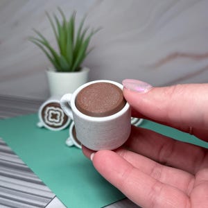 May include: A miniature white ceramic coffee cup with a brown coffee-like substance inside. The cup is being held in a hand.