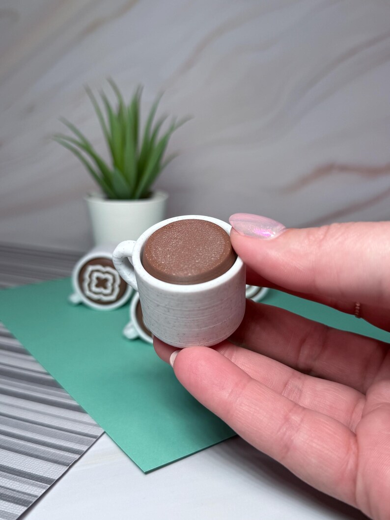 May include: A miniature white ceramic coffee cup with a brown coffee-like substance inside. The cup is being held in a hand.