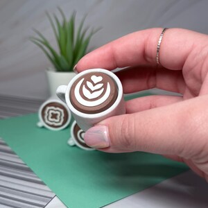 May include: A small white ceramic coffee cup with a brown liquid and a white latte art design. The cup is being held by a hand.