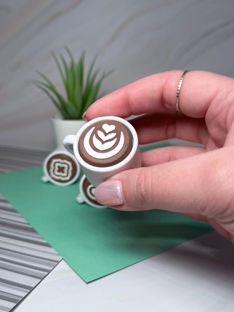 May include: A small white ceramic coffee cup with a brown liquid and a white latte art design. The cup is being held by a hand.