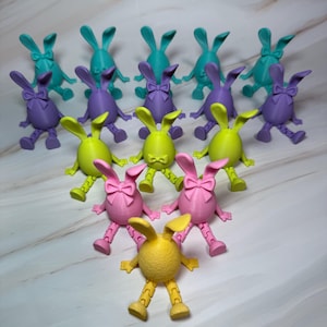 May include: A group of 12 colorful Easter bunny figurines, made of 3D printed plastic. The bunnies are arranged in a circle, with the yellow bunny in the center. The bunnies are all different colors, including pink, green, purple, and blue.