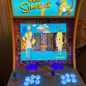 Puede incluir: Una máquina recreativa de The Simpsons con un esquema de color amarillo y azul. La pantalla muestra los personajes de Los Simpson y el texto "The Simpsons Arcade". La máquina tiene joysticks y botones.