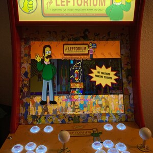 Puede incluir: Una máquina recreativa amarilla y roja con el texto "The Leftorium" y personajes de dibujos animados. La máquina tiene múltiples botones y joysticks, con una pantalla que muestra varios gráficos y texto.