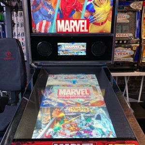 Puede incluir: Una máquina de pinball de Marvel con una superficie de juego de vidrio y una pantalla digital. La máquina tiene un gabinete negro y plateado con un logotipo de Marvel rojo y amarillo. La superficie de juego presenta una imagen colorida de personajes de Marvel, incluidos Spider-Man, Capitán América y Wolverine. La pantalla digital muestra el logotipo de Marvel y las palabras "Pinball".