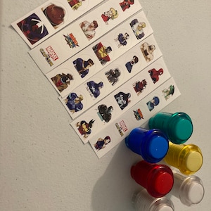 Puede incluir: Una colección de botones de juegos arcade en varios colores, incluyendo rojo, azul, verde, amarillo y transparente. También hay varias tiras de pegatinas con personajes de Marvel y Capcom, con los logotipos de Marvel y Capcom visibles.