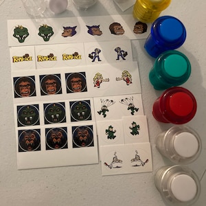Puede incluir: Un conjunto de botones de juego de arcade y pegatinas con varios diseños, incluyendo un monstruo verde, un lobo, un mono y un personaje con un paracaídas. Los botones son de varios colores, incluyendo amarillo, azul, verde y rojo. Las pegatinas están en hojas blancas.