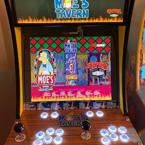 Puede incluir: Una máquina recreativa "Moe's Tavern" con acabado de madera. La máquina tiene una pantalla que muestra varias opciones de juego, incluyendo "Moe's Tavern" y "Tapper". El panel de control tiene joysticks y botones iluminados. La parte superior de la máquina muestra los logos "The Simpsons" y "Moe's Tavern".