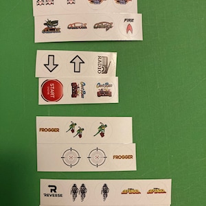 Puede incluir: Un conjunto de 16 pegatinas redondas con gráficos de videojuegos retro. Las pegatinas incluyen imágenes de naves espaciales, tanques, objetivos y otros elementos del juego. Las pegatinas están impresas en papel blanco con contornos negros.