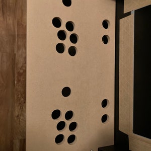 May include: A light brown wooden panel with multiple circular cutouts. The panel is attached to a black surface with screws.