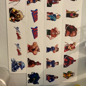 Puede incluir: Un conjunto de 18 pegatinas redondas con personajes de los videojuegos X-Men y Street Fighter. Las pegatinas son blancas con imágenes coloridas de los personajes. Las pegatinas están dispuestas en tres filas de seis pegatinas cada una.