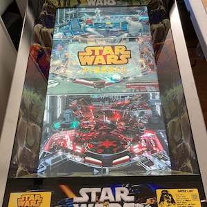 May include: A Star Wars pinball machine with a black and silver cabinet. The game features a digital display with images of the Star Wars characters and a game list. The machine is set to free play.