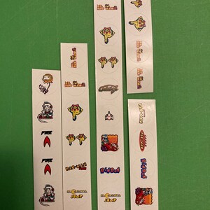 Puede incluir: Una hoja de pegatinas con personajes de videojuegos clásicos como Pac-Man, Dig Dug y Galaga. Las pegatinas son redondas y presentan ilustraciones coloridas de los personajes.