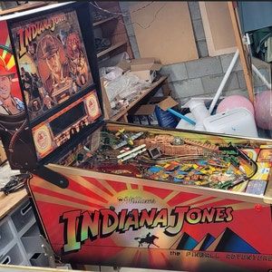May include: A vintage pinball machine with a red, gold, and black cabinet. The machine is titled "Indiana Jones: The Pinball Adventure". The cabinet features a graphic of Indiana Jones with a whip and a fedora.