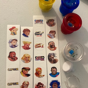 Puede incluir: Tres juegos de botones de joystick de arcade con tapas de diferentes colores: amarillo, azul y rojo. Cada juego incluye un botón, un anillo de plástico transparente y un botón de plástico blanco. Los botones están decorados con pegatinas con personajes del videojuego Street Fighter II.