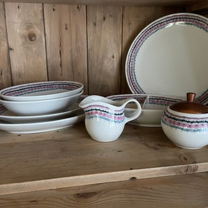 May include: A set of white ceramic dishes with a pink and gray floral pattern. The set includes a large dinner plate, a smaller bowl, a creamer, and a sugar bowl with a wooden lid.