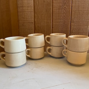 May include: Eight beige ceramic coffee mugs stacked in pairs on a white surface. The mugs have a simple design and are perfect for everyday use.