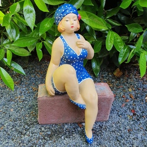Könnte beinhalten: Eine Vintage-Figur einer Frau in einem blauen Badeanzug mit weißen Sternmustern, passender Mütze und Schuhen. Die Figur sitzt auf einem roten Ziegelstein, mit einer roten Blume im Haar. Der Hintergrund zeigt grünes Laub und kleine graue Steine.