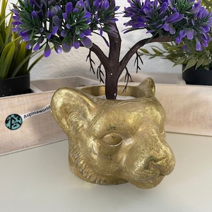 Puede incluir: Un macetero dorado con forma de cabeza de tigre con una pequeña planta morada en su interior.