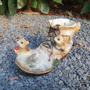 Puede incluir: Un macetero de bota de cerámica blanca con dos figuritas de ardillas. La bota tiene un cordón marrón y un detalle de musgo verde. Las ardillas sostienen una baya roja y una nuez.