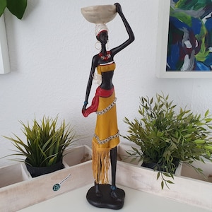 Puede incluir: Una figura negra de una mujer que lleva un cuenco en la cabeza. Lleva una falda amarilla con una faja roja y joyas de cuentas. La figura está de pie sobre una base negra.