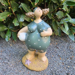 Könnte beinhalten: Keramikfigur einer Frau in einem grünen Badeanzug mit weißen Punkten, die einen weißen Ball hält. Die Figur steht auf einem sandigen Sockel mit einem lila Seestern. Die Frau hat braune Haare und trägt eine passende Badekappe.
